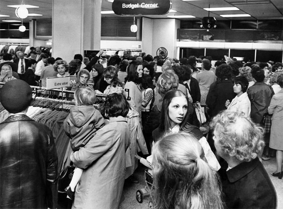 Black and white photo of a crowded department store, with many women shopping around clothing racks. The "Budget Corner" sign hangs from the ceiling above the busy crowd. Several shoppers are talking or browsing.