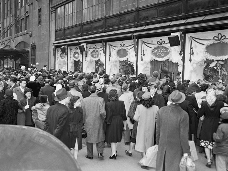 A large crowd of people in coats and hats gathers outside a department store with holiday decorations, peering into festive window displays labeled "Santa Claus Soon.