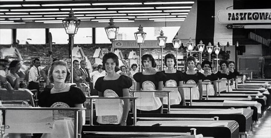 A row of female cashiers in matching uniforms stand behind checkout counters in a busy, brightly-lit supermarket with signs and shoppers in the background.