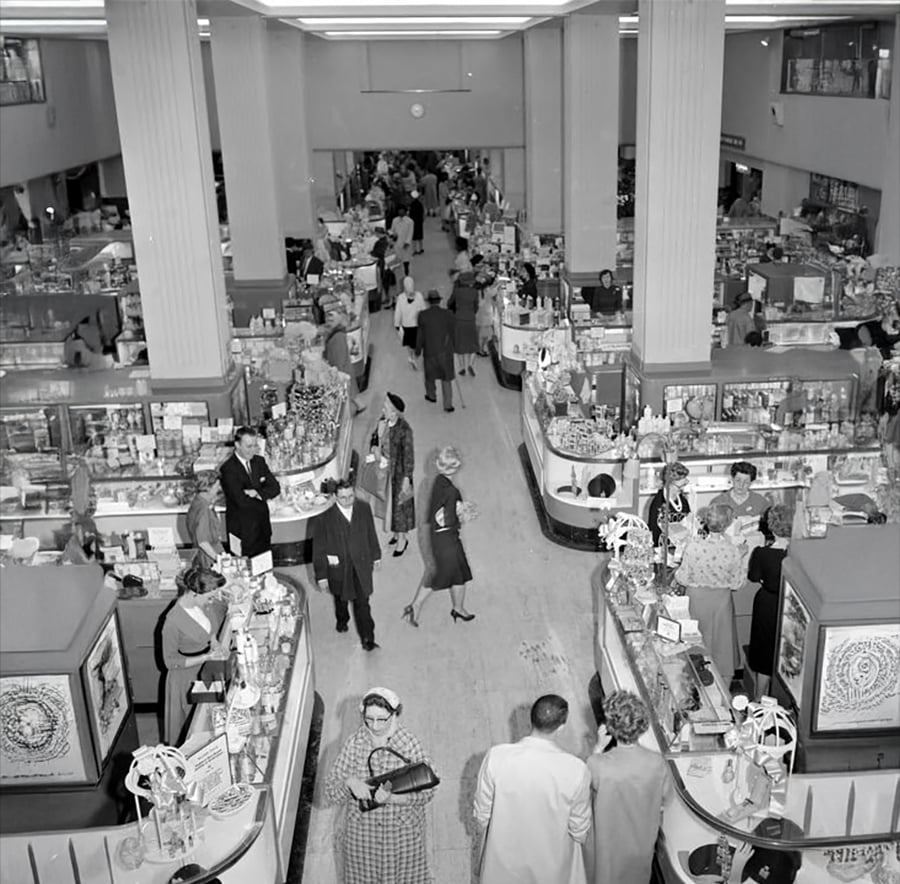 Black and white photo of a busy department store in the mid-20th century, with shoppers browsing and clerks assisting at counters filled with various merchandise under bright overhead lights.