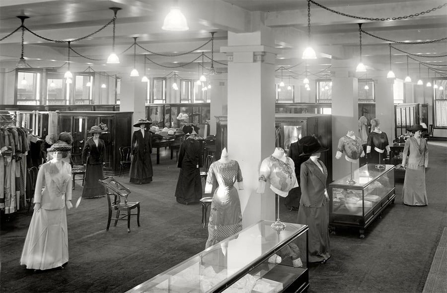 Early 1900s department store with mannequins displaying women's fashion, glass display cases, and customers browsing clothes and hats under hanging lights in a spacious, elegant interior.