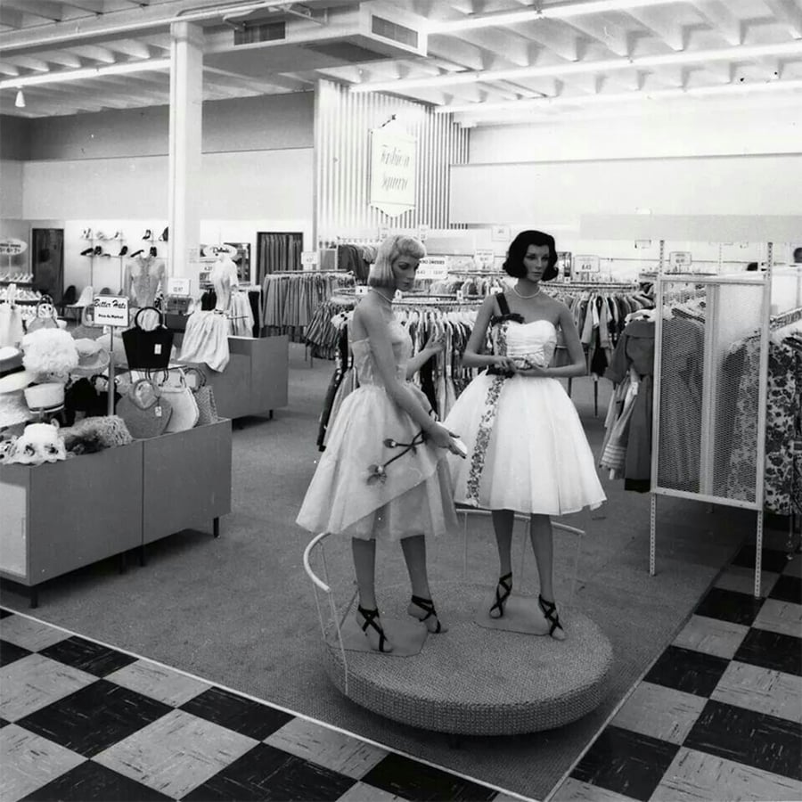 Two mannequins in vintage dresses stand on a small platform in a mid-century clothing store, surrounded by racks of clothes and accessories under bright ceiling lights.