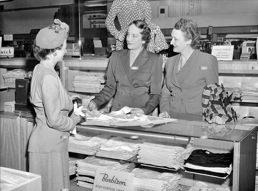 A woman in a hat talks to two saleswomen behind a store counter stacked with folded clothes. One saleswoman holds a garment while the other stands beside her. The setting appears to be a mid-20th-century clothing store.