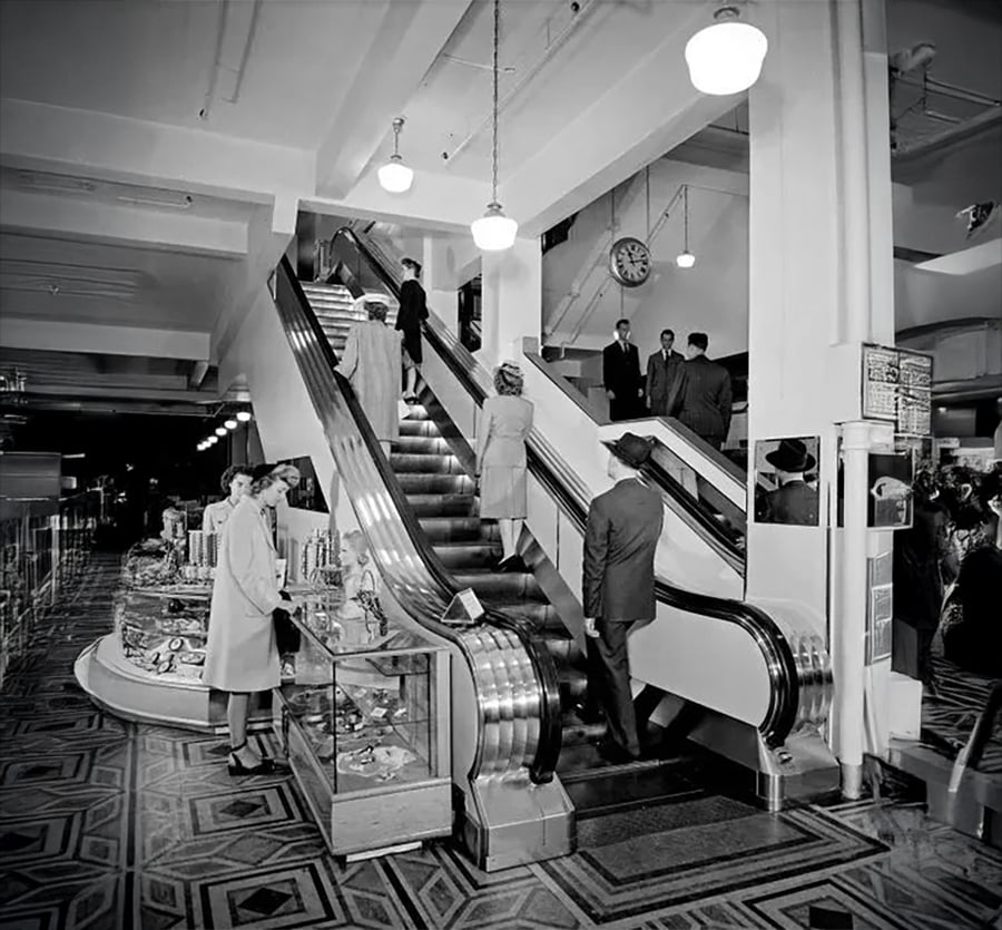 Black and white photo of people in 1940s-style clothing using an escalator in a department store, with display cases and a clock visible in the background.