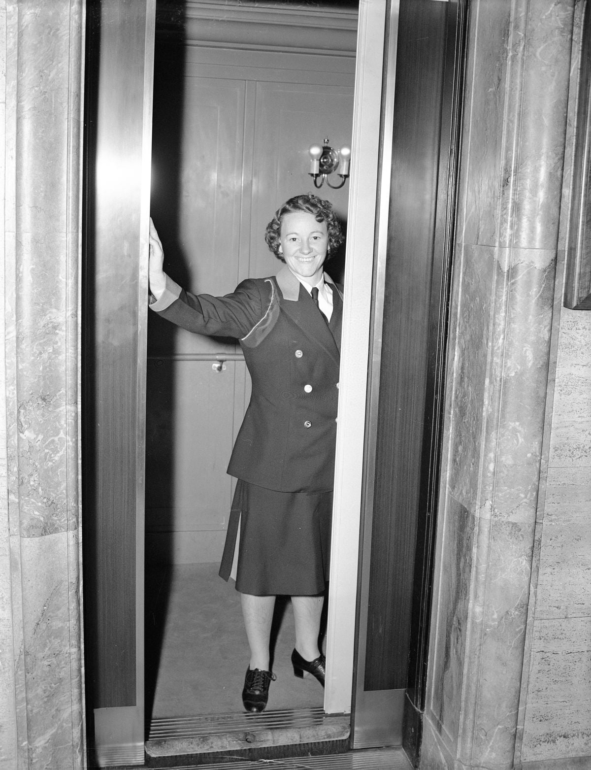 A woman in a uniform stands in the doorway of a partially open elevator, smiling at the camera. The setting appears to be indoors, with marble walls and a light fixture visible behind her.