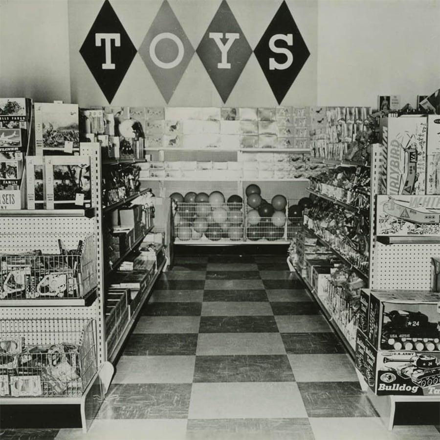 Black-and-white photo of a toy store aisle with shelves full of toys on both sides. The floor is checkered, and a wall at the end displays the word "TOYS" above bins of balls and other items.