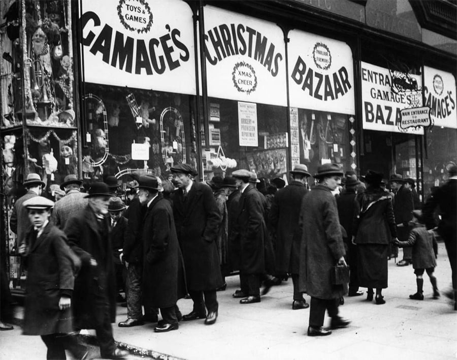 A crowd of people in winter coats and hats stands outside a store decorated with Christmas signs reading "Gamages Christmas Bazaar." Shop windows display toys and gifts, and festive decorations are visible inside.
