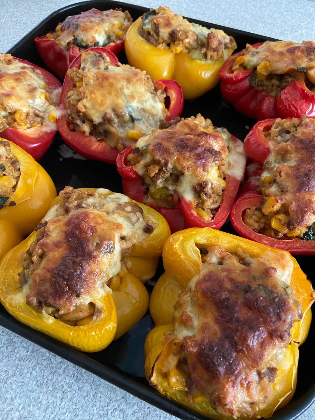 A tray of baked stuffed bell peppers, both red and yellow, filled with a mixture of vegetables, grains, and topped with melted, golden-brown cheese.
