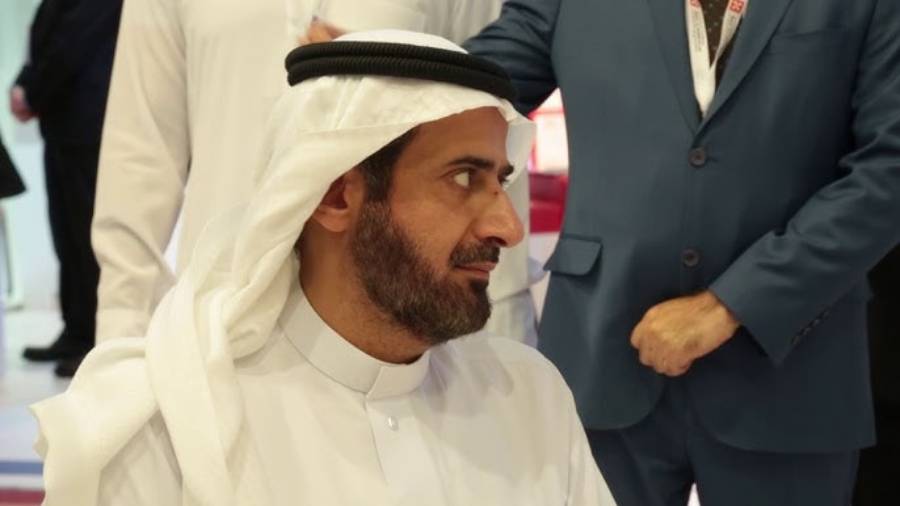 A man in traditional Middle Eastern attire, including a white kandura and a white headscarf with black agal, sits and looks to the side while others in formal clothing stand in the background.