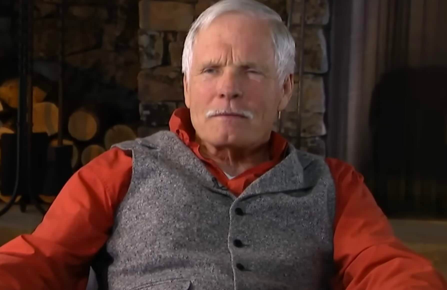 An older man with white hair and a mustache sits indoors, wearing a red shirt and a grey vest. There is a stone wall and stacked firewood in the background.