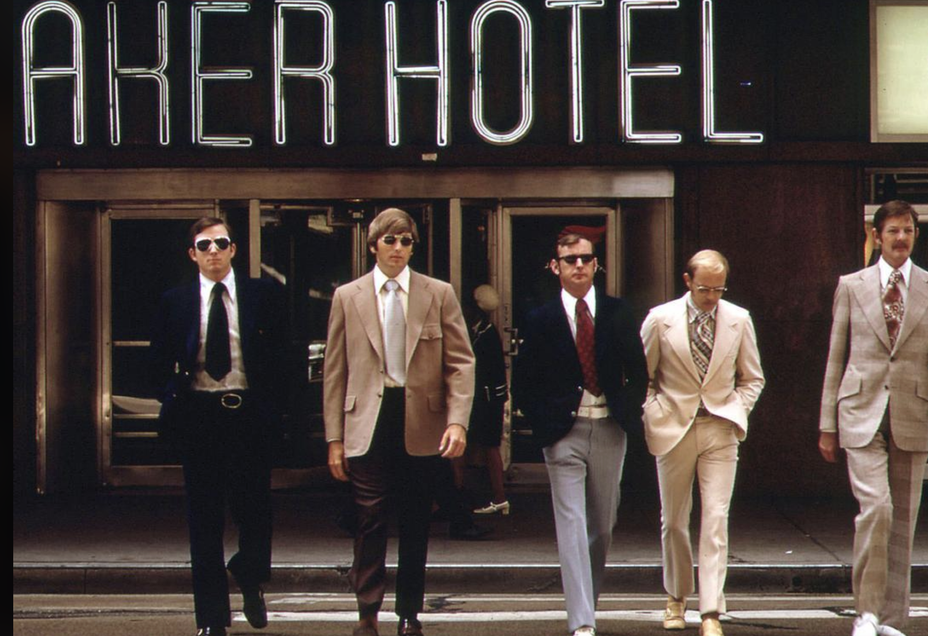 Five men in suits and sunglasses walk confidently across a street in front of a building with a large neon sign reading "AKER HOTEL." The scene appears stylish and retro, suggesting the 1970s.