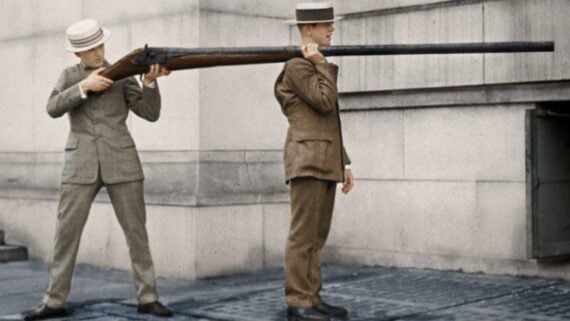 Two men in early 20th-century suits and straw hats pose with an extremely long gun; one aims while the other stands holding the barrel on his shoulder near a stone building.