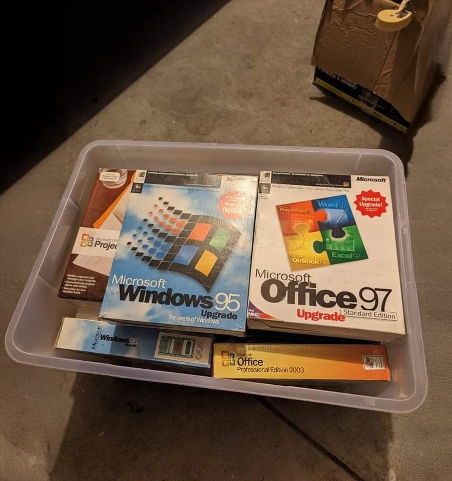 A plastic bin filled with vintage Microsoft software boxes, including Windows 95, Office 97, Project, and Office Professional Edition 2003, sitting on a gray floor.