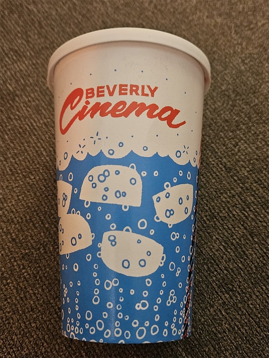 A blue and white disposable cup with the words "Beverly Cinema" in red and white, decorated with cartoon bubbles and floating shapes, placed on a brown textured surface.