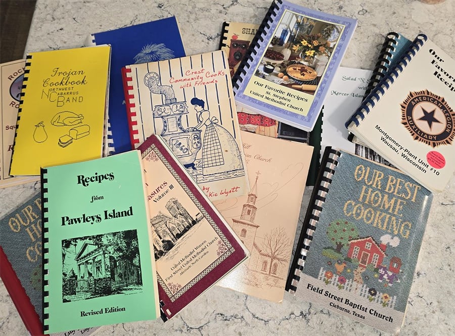A collection of various spiral-bound community and church cookbooks with colorful covers is spread out on a marble countertop. Each cover features different illustrations, text, and designs.