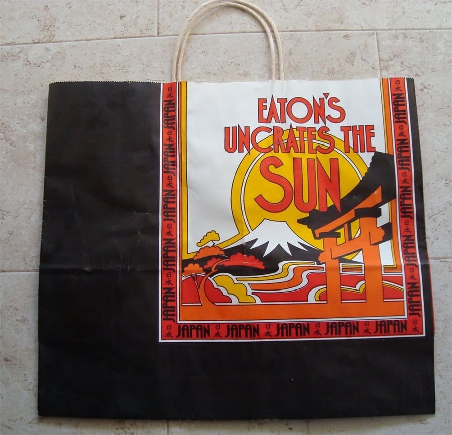 A paper shopping bag with a design showing Mount Fuji, a rising sun, and a torii gate. Text reads “Eaton’s Uncrates the Sun.” The border and bottom edge feature repeated "Japan" lettering.