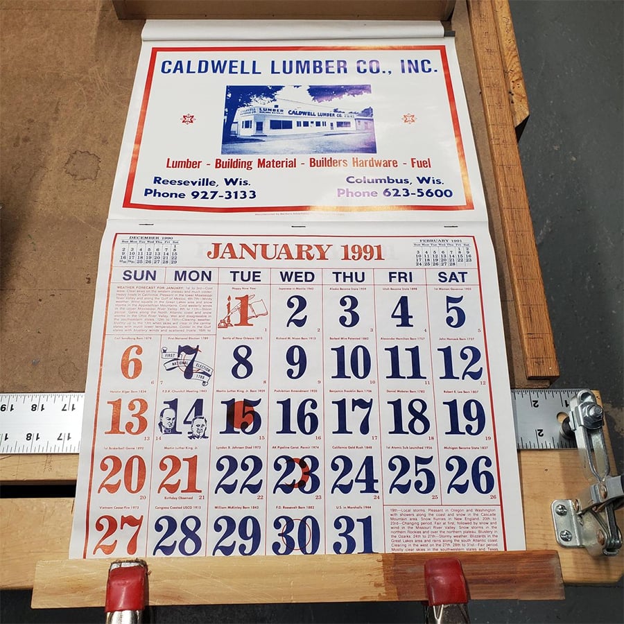 A vintage calendar showing January 1991 with dates in bold red and blue numbers, above an ad for Caldwell Lumber Co., Inc., displayed on a wooden surface with red metal clips holding it open.