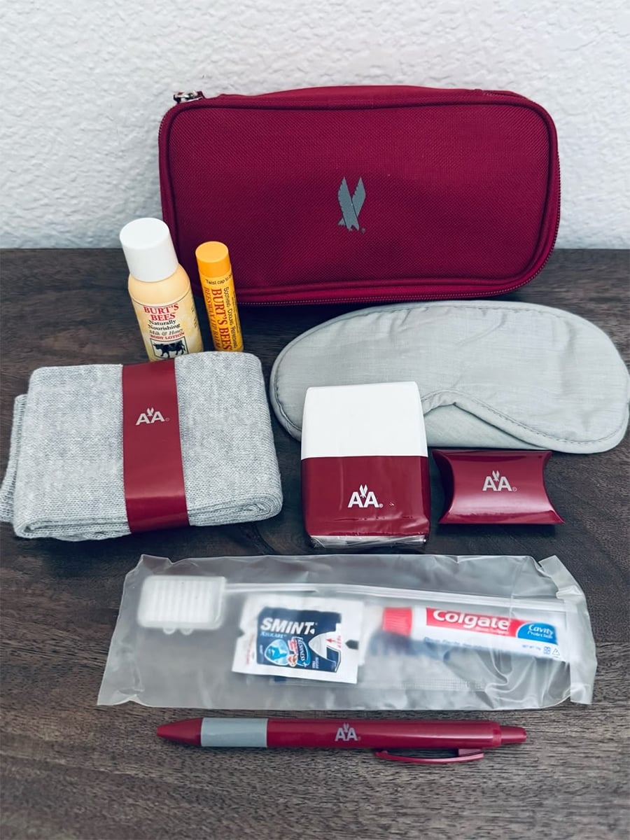 A burgundy American Airlines amenity kit with a gray eye mask, gray socks, Burt’s Bees lotion and lip balm, a dental kit with Colgate toothpaste, Smint mints, a pen, and two small boxes, all arranged on a wooden surface.