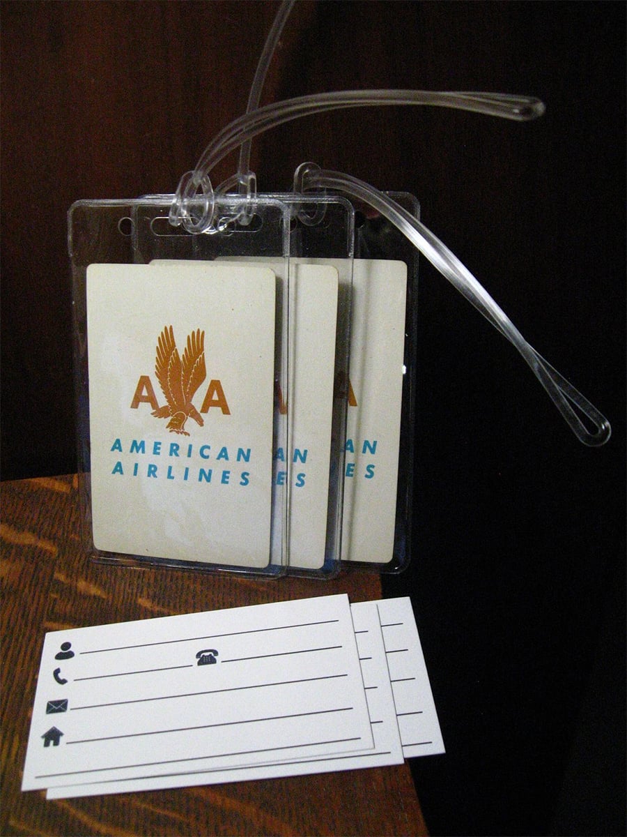 Three American Airlines luggage tags with eagle logos and clear plastic straps are hanging together. Two blank tags with contact information icons and lines are placed below them on a wooden surface.