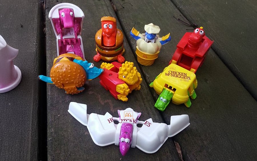 A group of colorful McDonald’s Happy Meal McNugget toys, each dressed in different costumes, is arranged on a wooden surface. The toys are shaped like chicken nuggets with cartoonish faces.