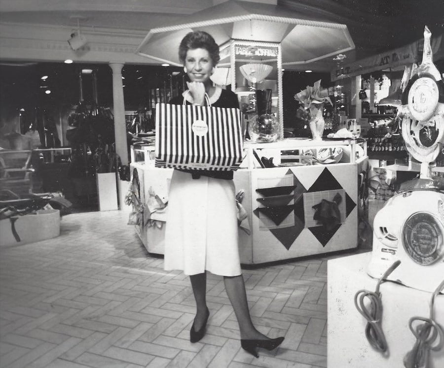 A woman stands smiling in a department store, holding a large striped shopping bag. She wears a skirt and blazer, and the store displays various accessories and decorative items in the background. The image is black and white.