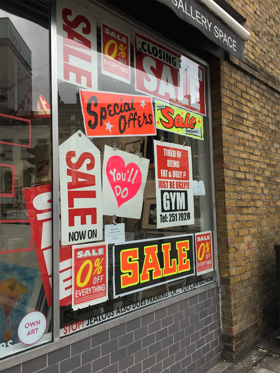 A shop window covered with colorful sale signs, special offers, and discount posters. One sign advertises a gym with the tagline “Tired of being fat & ugly? Just be ugly!” The shop exterior is brick.