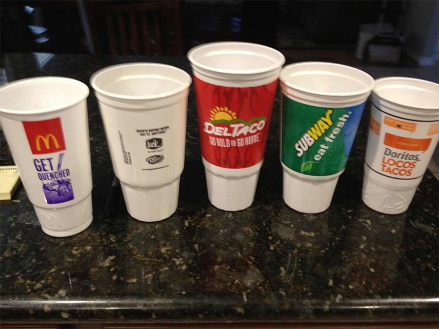 Five large fast food cups from McDonald’s, Taco Bell, Del Taco, Subway, and Doritos Locos Tacos are lined up on a dark kitchen counter. Each cup displays its brand’s logo and colors.