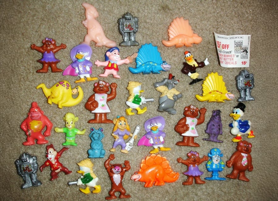 A collection of colorful plastic toy figures, including animals, cartoon characters, dinosaurs, and robots, is arranged in rows on a brown carpet. A coupon and several duplicates are visible among the toys.