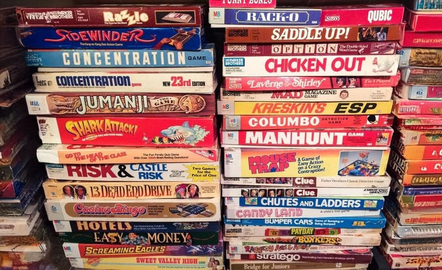 Stacks of vintage board games piled closely together, featuring colorful boxes with titles like Shark Attack, Risk, Clue, Chutes and Ladders, Candy Land, Life, and more.