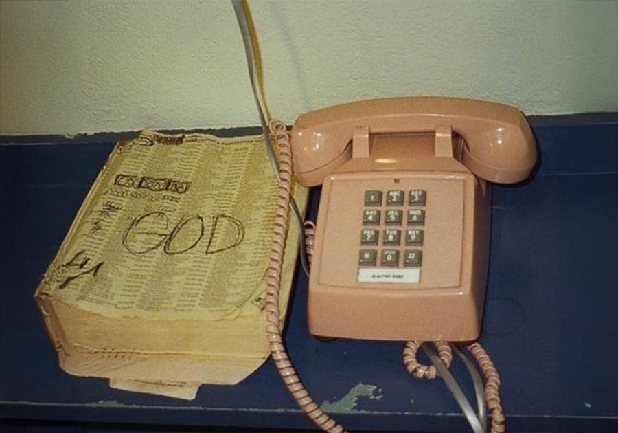 A worn yellow phone book with "GOD" written on the cover sits next to a vintage pink landline telephone on a blue tabletop.
