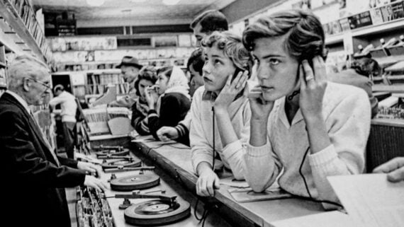 Black and white photo of young people in a record store, listening to vinyl records on headphones at a listening bar, with shelves of albums in the background and a clerk assisting at the counter.