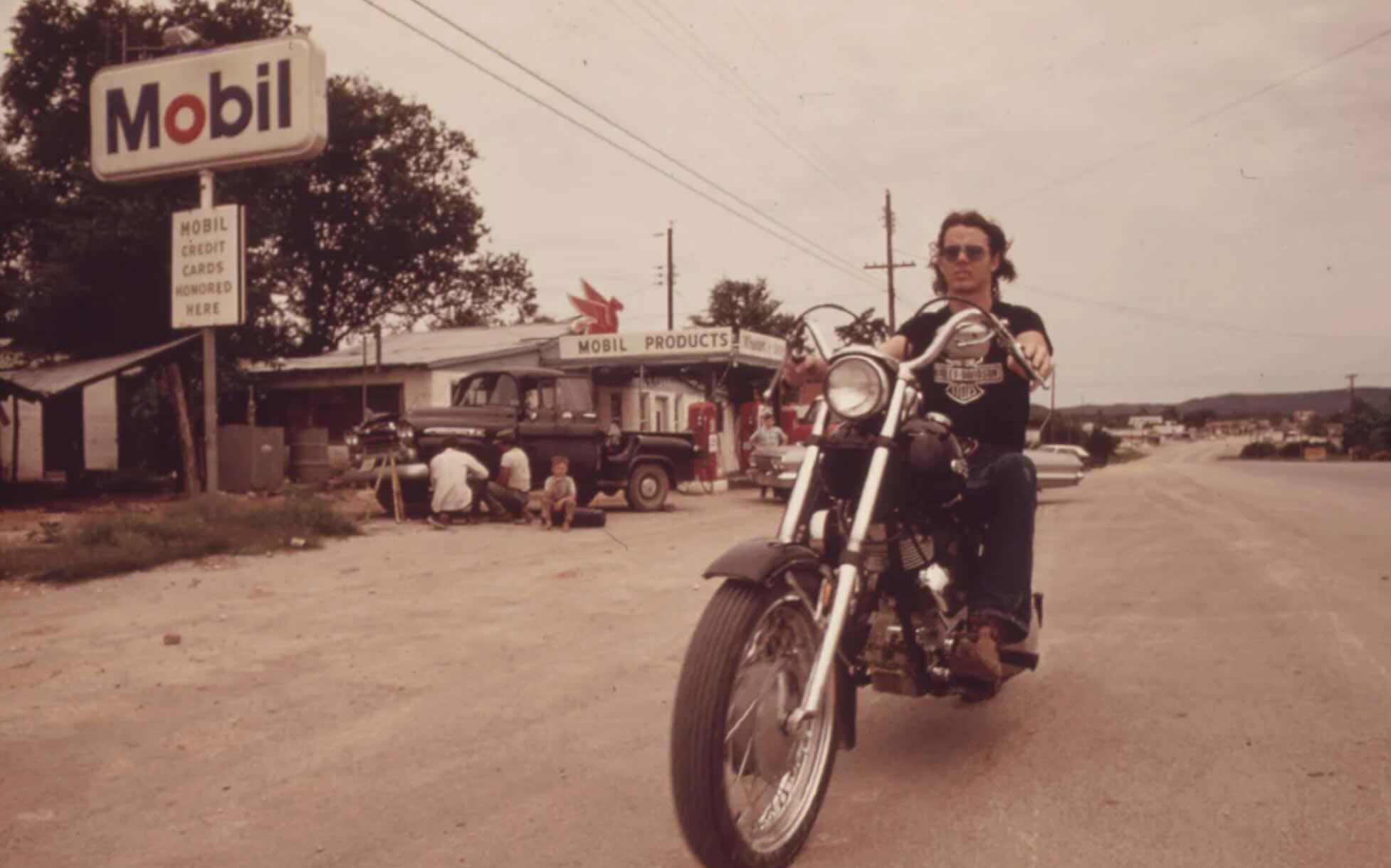 A person wearing sunglasses rides a motorcycle on a road near a Mobil gas station. Several people and a dog are near a vintage truck in the background. The scene appears to be from the past, with retro signage and vehicles.