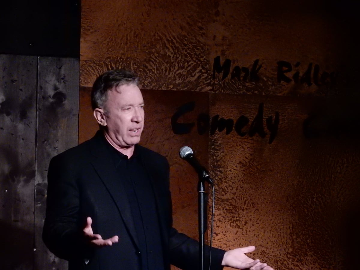 A man in a black suit stands on stage with a microphone, gesturing with his hands while performing at a comedy club. The background features a textured wall with partially visible text.
