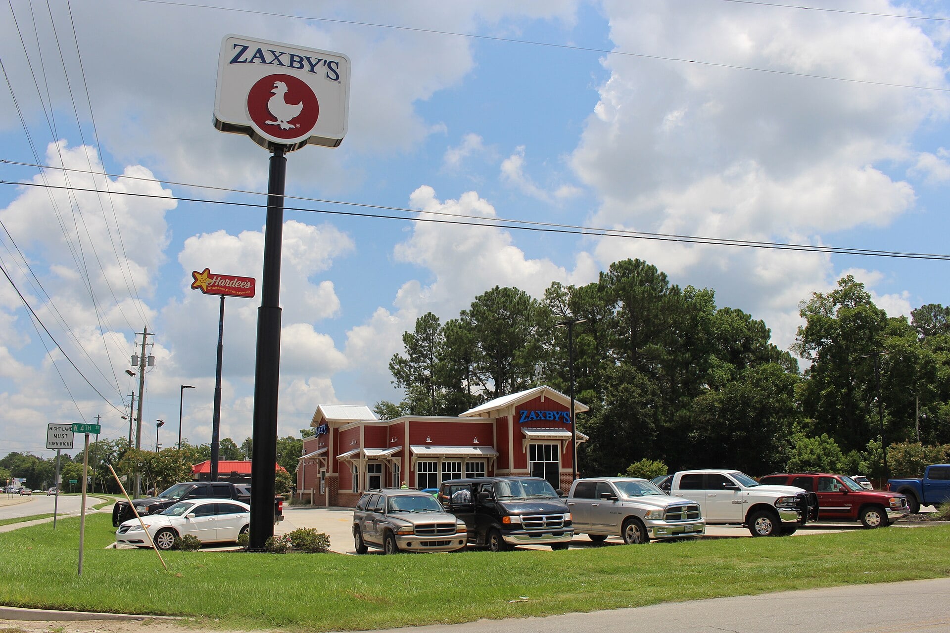 A Zaxby’s restaurant with several cars parked in front, a large Zaxby’s sign on a pole, and trees and clouds in the background on a sunny day.