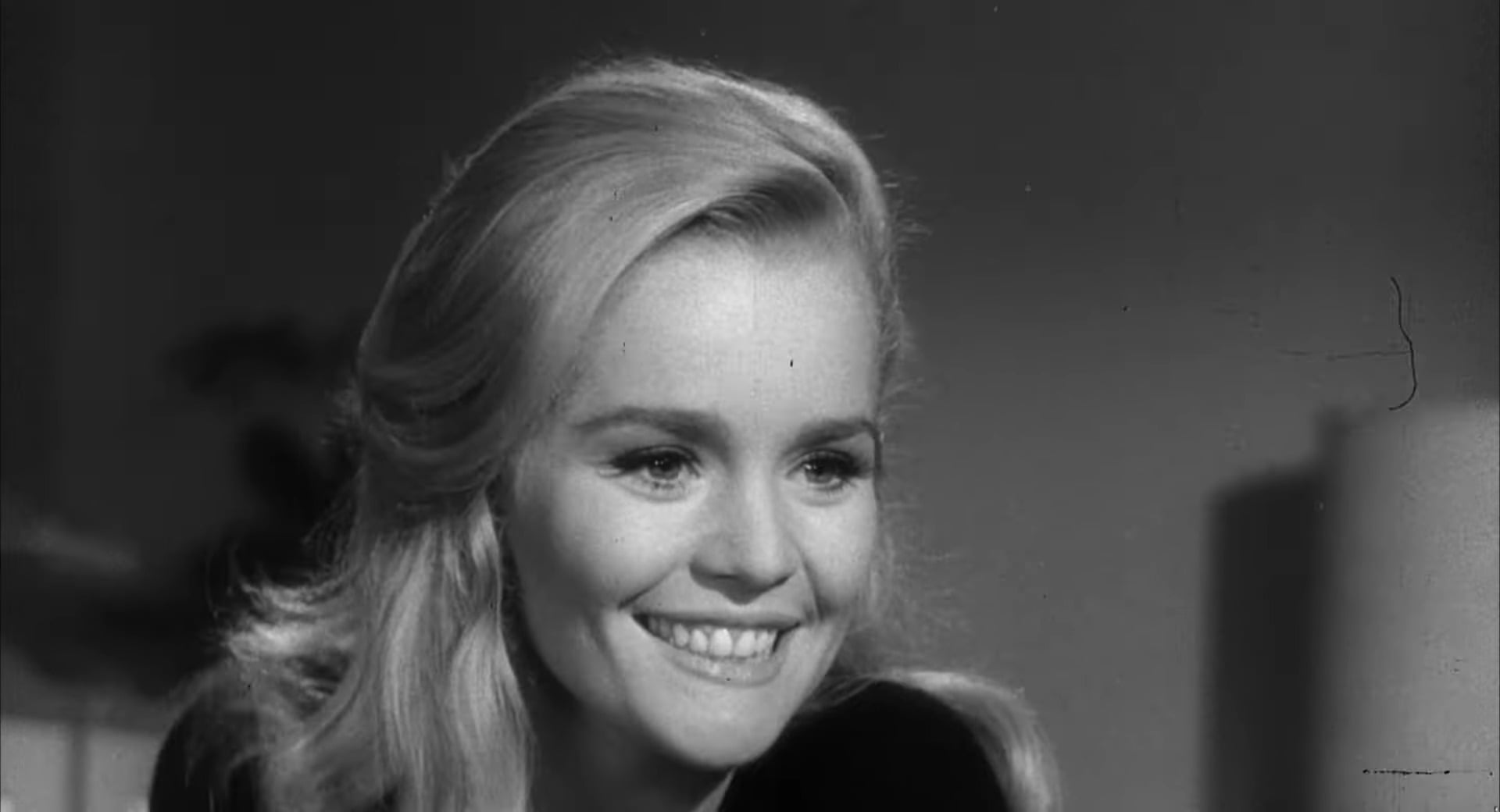A smiling woman with long, light-colored hair is shown in a close-up, black-and-white photo. She appears happy and is looking slightly off camera. The background is softly blurred.