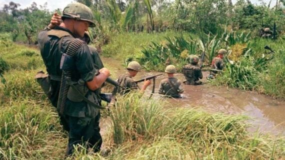 Five soldiers in green uniforms and helmets move through a swampy, grassy area, with weapons ready. Four soldiers wade through muddy water while one stands on the bank, watching the others. Lush vegetation surrounds them.