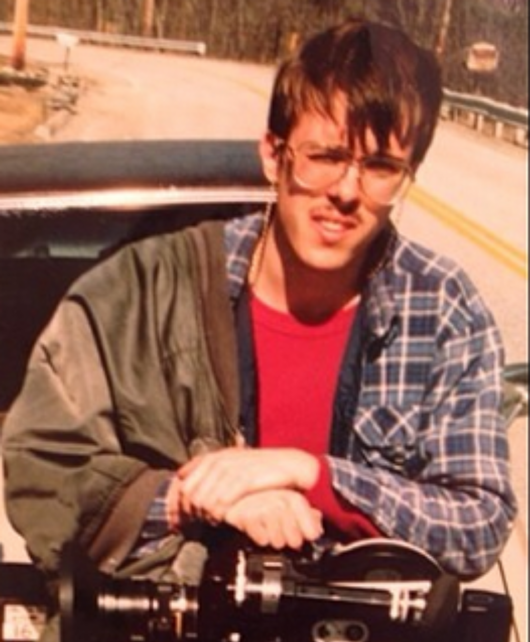 A young person in glasses and a plaid shirt stands outdoors, leaning on equipment mounted to a car, with a road and trees visible in the background.