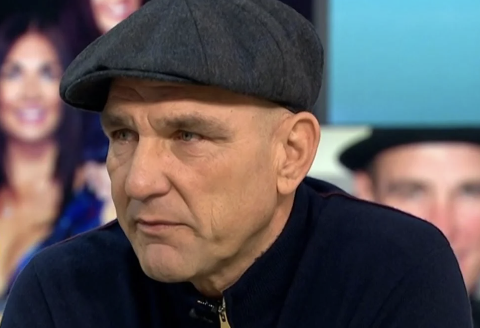 A middle-aged man wearing a flat cap and a dark zip-up sweater sits indoors, looking slightly to the side with a serious expression. The background is blurred with mostly blue and flesh tones.