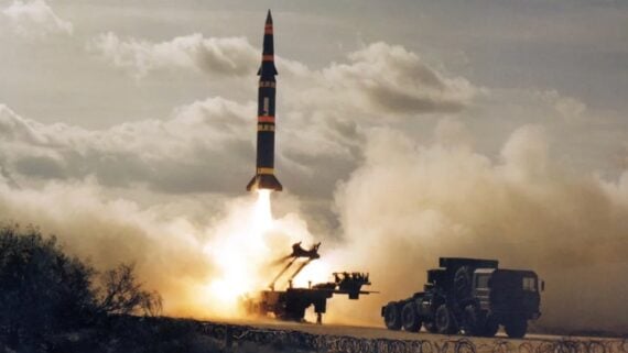 A missile launches vertically from a military vehicle-mounted platform, surrounded by smoke and dust, with cloudy skies and scattered vegetation in the background.