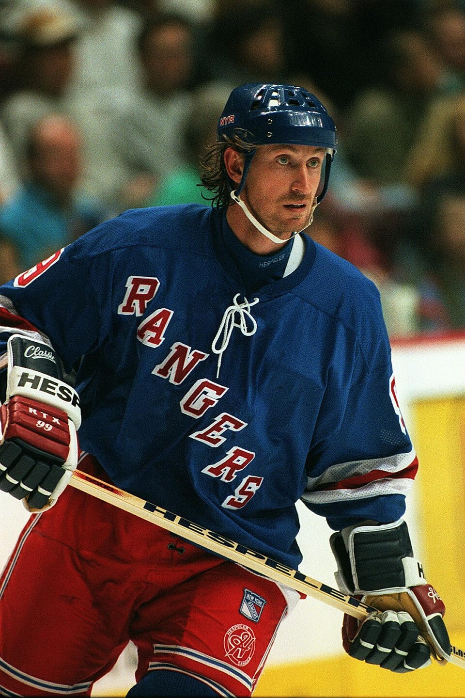 A hockey player in a blue New York Rangers jersey and helmet skates on the ice, holding a hockey stick, with blurred spectators in the background.
