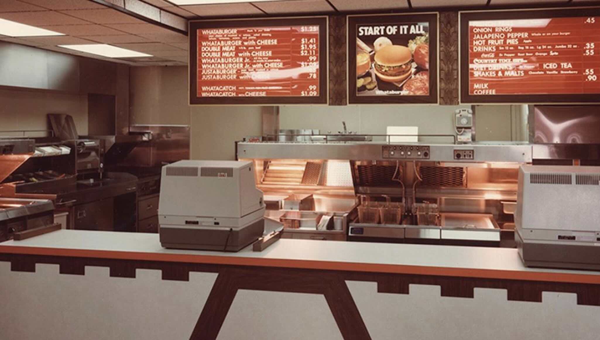 Vintage fast-food restaurant counter with two cash registers, overhead menu boards listing burgers and drinks, and a lit kitchen area in the background. The decor features brown, orange, and cream tones.