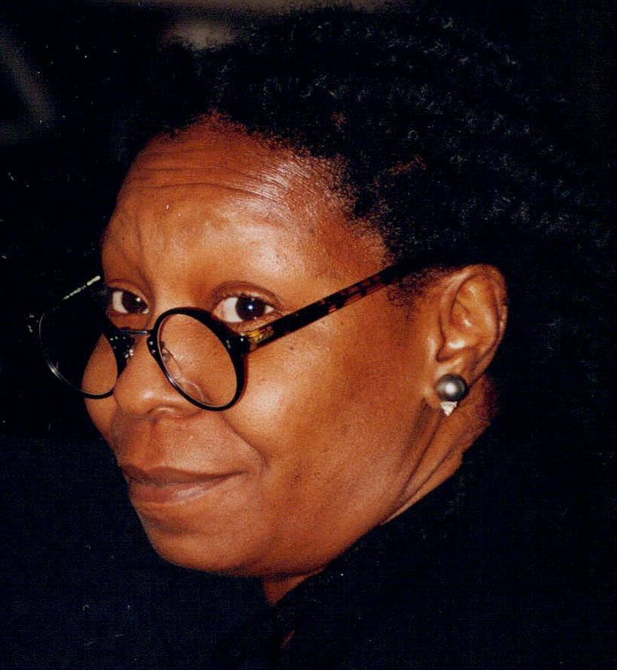 A person with short curly hair, wearing round tortoiseshell glasses and pearl earrings, looks toward the camera with a slight smile.