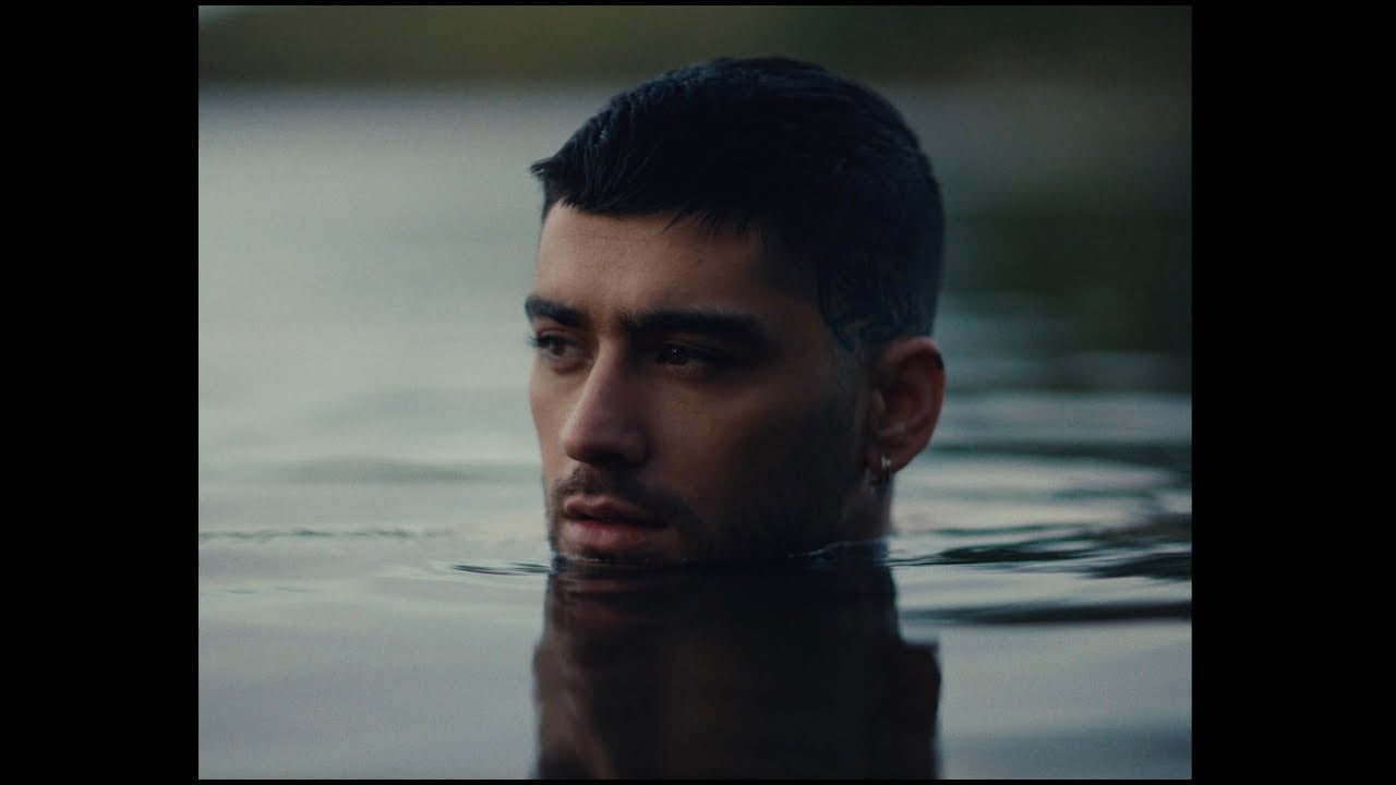 A man with short dark hair and a trimmed beard is partially submerged in calm water, with only his head visible above the surface. The background is blurred, creating a serene and contemplative mood.