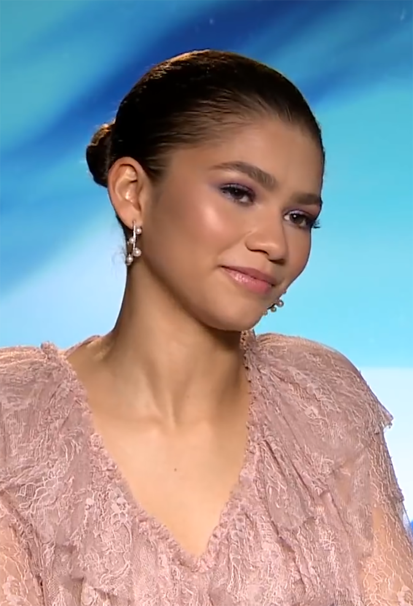 A woman with slicked-back hair in a bun, wearing pearl earrings and a light pink lace top, smiles slightly against a blue gradient background.