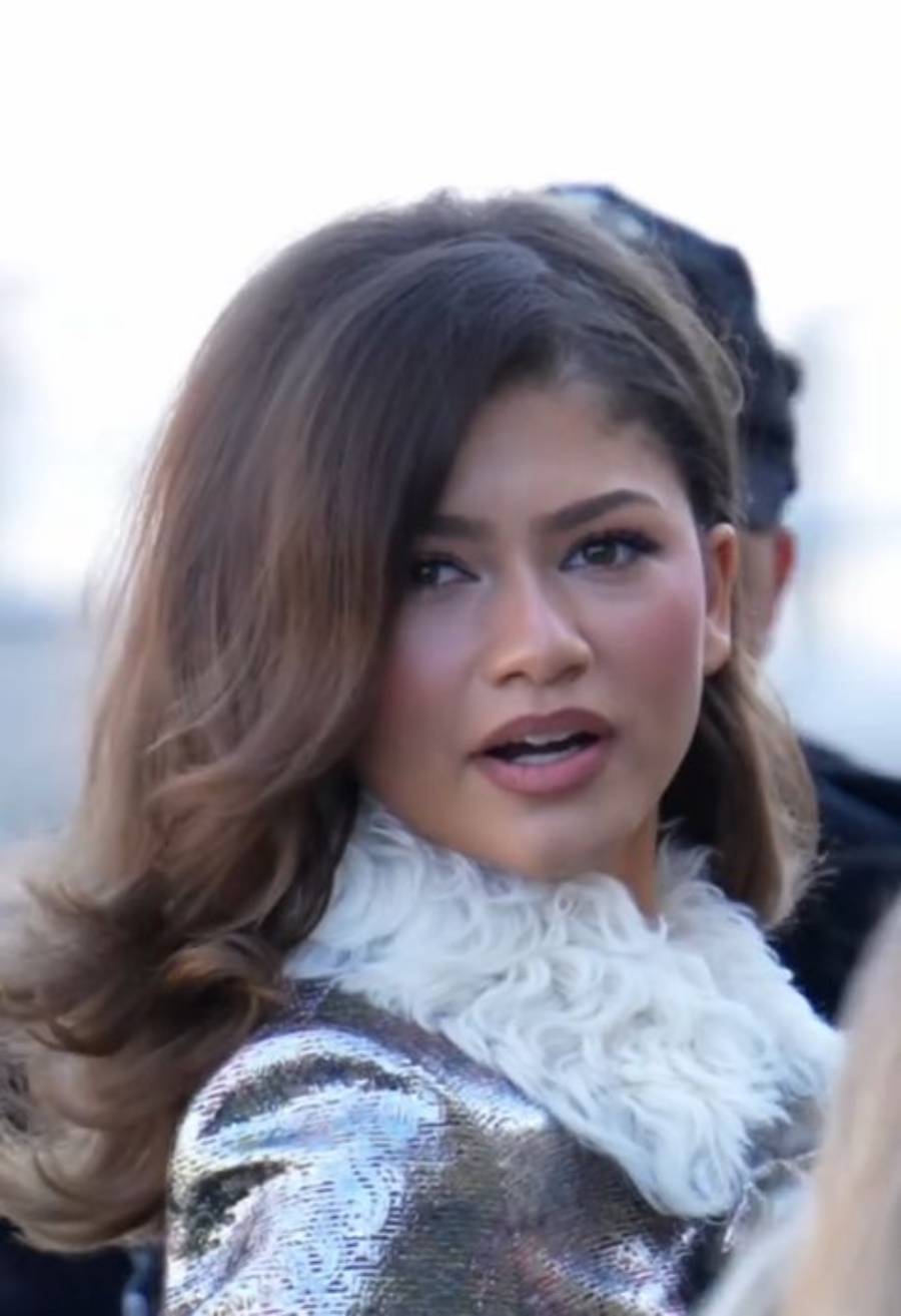 A young woman with wavy brown hair, wearing a shiny silver outfit with a white furry collar, looks slightly over her shoulder with her mouth slightly open. The background is softly blurred.