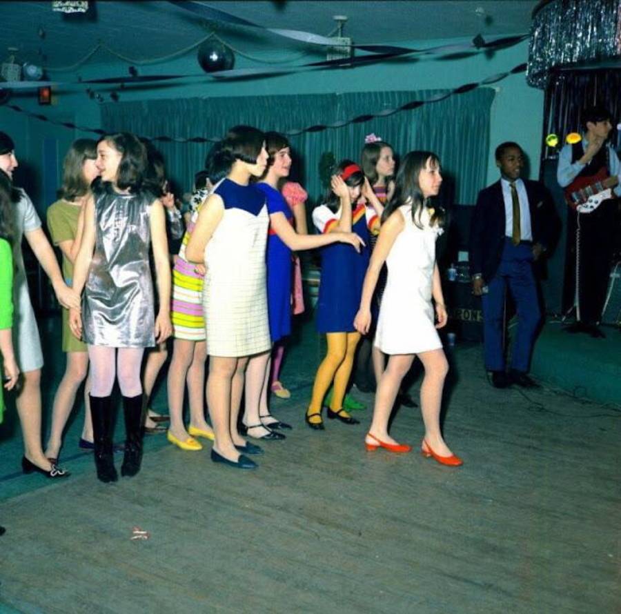 A group of young people in colorful 1960s-style clothing dance in a line on a wooden floor while musicians perform on a stage in the background. The room is decorated with curtains and a disco ball.