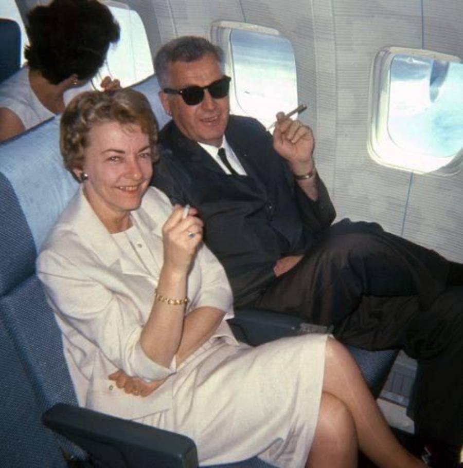 A woman and man sit in airplane seats, both smiling and holding cigarettes. The man wears sunglasses and a dark suit; the woman wears a light dress. Another passenger sits in front of them.