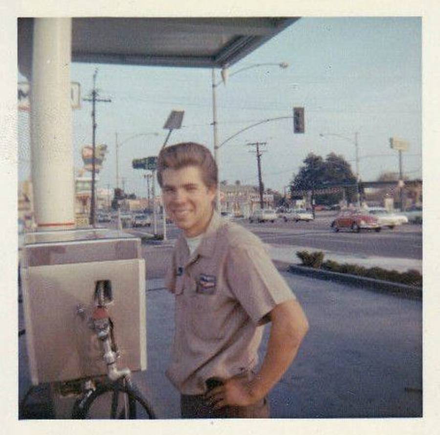 A young man in a work uniform smiles while standing next to a gas pump at a vintage gas station, with a street and cars visible in the background.