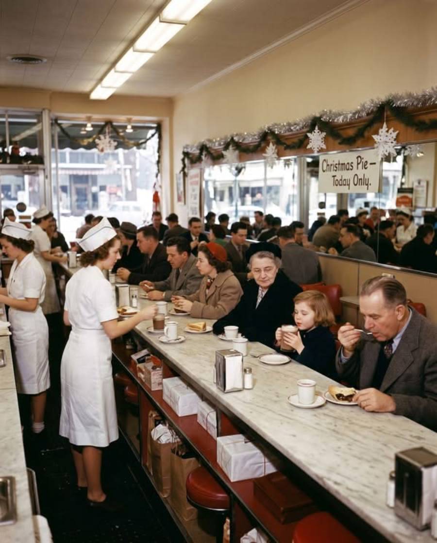 A busy 1950s diner with people eating at a long counter, served by waitresses in white uniforms; a “Christmas Pie - Today Only” sign hangs above.