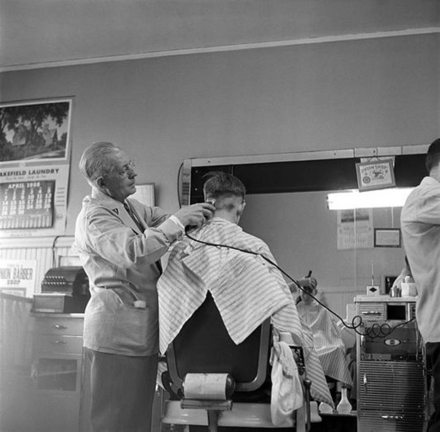 A barber in a white coat gives a haircut to a customer seated in a barber chair, both covered with striped capes. Mirrors, barber tools, and a wall calendar are visible in the black-and-white barbershop scene.